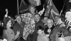 A jubilant crowd celebrates by lifting a smiling woman into the air. The scene is filled with waving flags and happy faces.