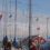 Sailboats with various flags docked at a marina. A red building with a peaked roof is in the background against a partly cloudy sky.
