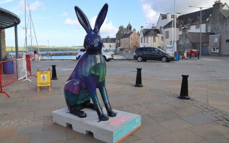 A colorful rabbit statue sits on a pedestal in a paved town square with buildings and cars in the background.