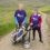 Two people wearing purple Alzheimer Scotland t-shirts stand on a golf course with a golf trolley and bags, surrounded by grassy hills and pathways.