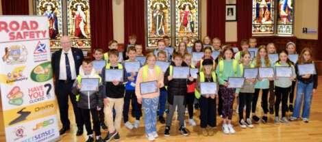 A group of children and an adult pose with certificates in front of a banner about road safety. Stained glass windows are in the background.