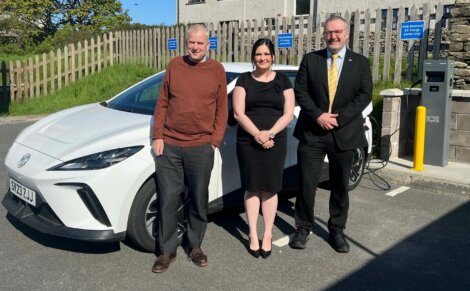 Three individuals stand in front of a white electric vehicle parked at a charging station. The man on the left wears a brown sweater, the woman in the center wears a black dress, and the man on the right wears a suit.