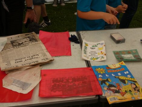 A table with various items including newspapers, comic books, red transparent sheets, and other papers. Two people are partially visible, engaged with the items.