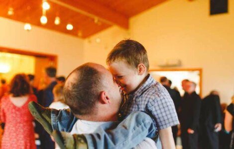 An adult man is holding a smiling boy in a room filled with people. The two are making eye contact, and the boy is wearing a checkered shirt and jeans.