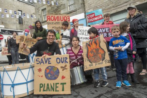 A group of people, including adults and children, hold signs advocating for environmental conservation. Signs include messages like "Save Our Planet," featuring a globe and flames.