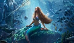 A mermaid with long flowing hair sits on a rock underwater surrounded by various sea creatures. An older man with a trident and a spectral figure look on in the background.