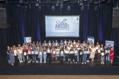 Group of people holding certificates on stage at the Saltire Awards Ceremony 2023, with a banner and screen displaying event information in the background.