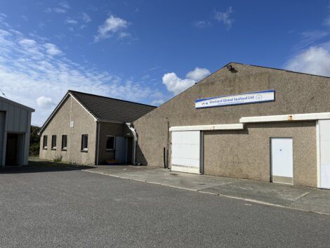 A concrete building with a sign reading "Shetland Global Seafood Ltd," featuring a large garage door and a smaller entrance, under a partly cloudy sky.