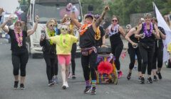 A group of people in colorful outfits and leis happily marching and dancing on a street during a parade. A truck is visible in the background.
