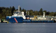 A large blue coastguard vessel docked at a pier with a tree-covered hillside in the background. The ship has a white and red striped emblem and various equipment on deck.