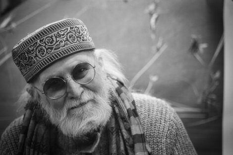 Black and white photo of an elderly man wearing round glasses, a patterned cap, and a scarf. He has a white beard and mustache and is looking at the camera.