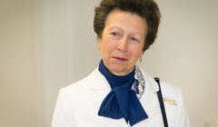 A woman with short, styled hair wears a white blazer, blue scarf, and a pin. She has a thoughtful expression and stands before a neutral background.
