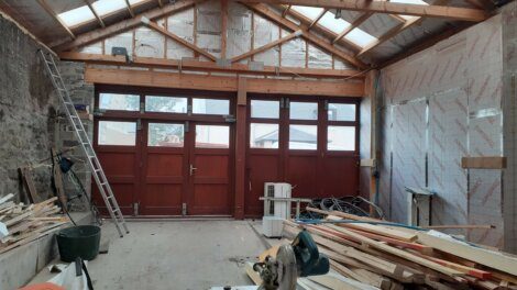 A partially renovated room with exposed insulation, red doors, a ladder, and construction materials scattered around. Skylights are present in the ceiling.