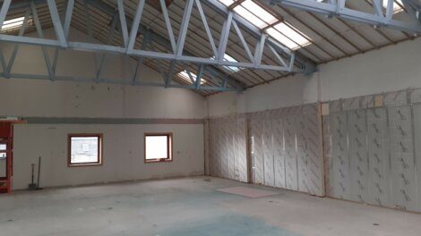 A large, empty room with a high ceiling and exposed trusses. There are two windows, one of which is partially covered by insulation on the wall. The floor appears unfinished.