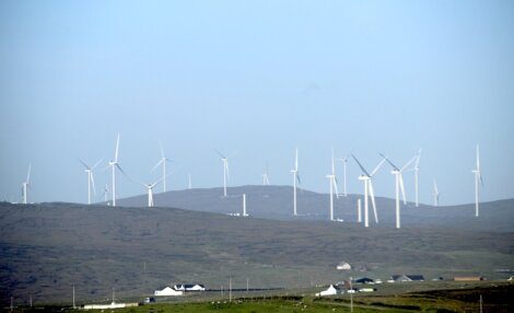 Several wind turbines spread across a hilly landscape under a clear sky, with a few buildings visible in the foreground.