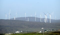 Several wind turbines spread across a hilly landscape under a clear sky, with a few buildings visible in the foreground.
