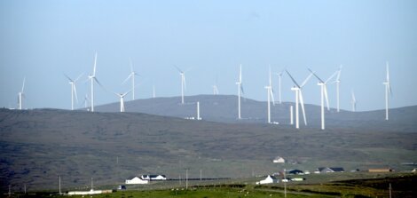 Several wind turbines spread across a hilly landscape under a clear sky, with a few buildings visible in the foreground.