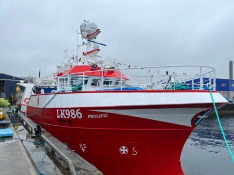 A red and white fishing vessel docked beside a pier with the identification "LX986 Prolific." Various equipment is visible on the boat, and colorful flags are strung along the mast.