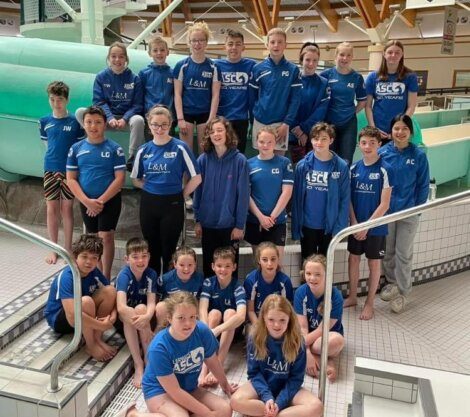 A group of young swimmers wearing blue uniforms poses for a team photo at an indoor pool facility. Some are standing while others sit on the edge of the pool area.
