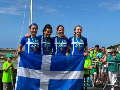 Four athletes in blue and white uniforms hold a blue and white flag, displaying medals around their necks. They stand outdoors with spectators in the background.