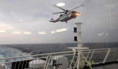 A red and white helicopter hovers above a ship's deck at sea with cloudy skies in the background.