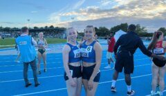 Two female athletes wearing blue athletic uniforms stand on a blue track, smiling at the camera. Other people and athletes are in the background, along with a cloudy sky at sunset.