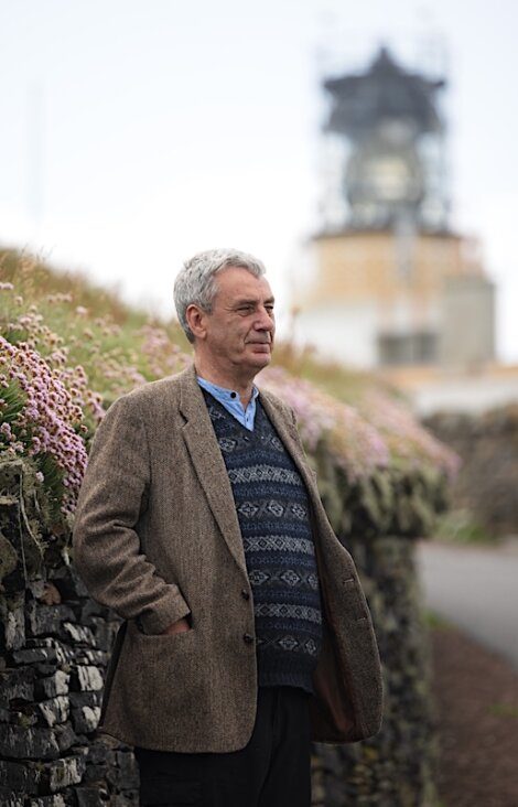 A man in a tweed jacket stands with hands in pockets beside a stone wall with blooming flowers, with a blurred lighthouse in the background.