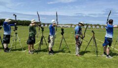 A group of people standing on a grassy field, aiming recurve bows at targets set up in the distance under a clear blue sky. Tripods are placed beside some archers.