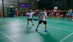 Indoor badminton game in progress. A player in a white shirt prepares to hit the shuttlecock while another player in a blue shirt and black shorts stands ready. Spectators watch from the sideline.
