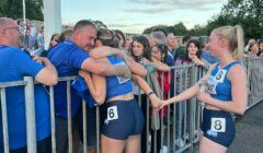 Athletes wearing competition numbers 6 and 8 stand next to a metal barrier; number 6 hugs a man, while number 8 holds hands with number 6. Spectators watch from the other side of the barrier.