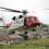 A red and white Coastguard rescue helicopter is in flight above a residential area with houses in the background.