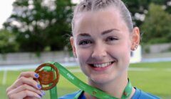 A person with braided hair and a blue shirt holds up a gold medal with a green ribbon, smiling. Trees and a track are visible in the background.