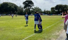 Three soccer players in blue jerseys, one with the number 12, celebrate near the sideline, while players from both teams are scattered around the field. Spectators watch from the sides on a partly cloudy day.