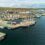 Aerial view of a coastal town featuring docks with boats and ships, colorful buildings, and rolling hills in the background.