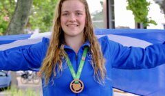 A person with long wet hair smiles while holding out a blue and white flag. They are wearing a blue jacket and a green lanyard with a bronze medal around their neck. Trees and a building are in the background.