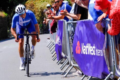 Cyclist in blue racing uphill past a cheering crowd behind barriers with NatWest branding.