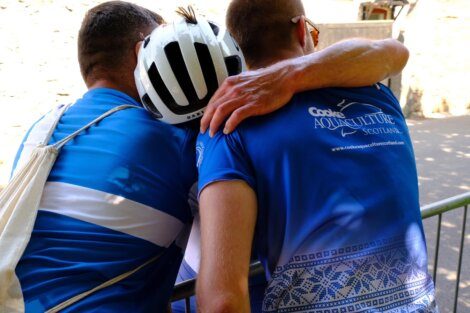 Three cyclists in blue jerseys stand next to a metal barrier, with one cyclist wearing a helmet and another placing an arm around a fellow cyclist’s shoulder.