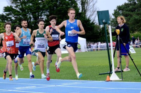 A group of male athletes running on a track, with one runner in a blue uniform leading the race. A woman is standing near the finish line, observing the race. Trees and spectators are in the background.