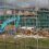 Construction site with a blue excavator, building material debris, and a partially completed green-clad building in the background, viewed through a chain-link fence on a cloudy day.