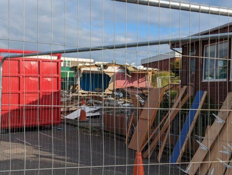 Construction site with demolished building debris behind a metal fence. Red shipping container on the left, and wooden planks and traffic cone in the foreground. Clear sky above.