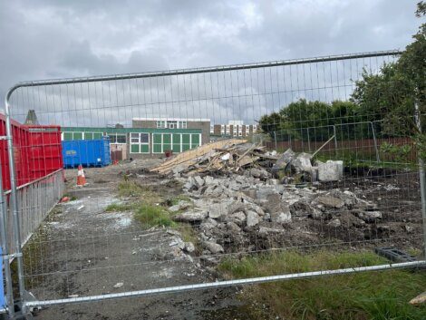 Construction site behind a metal fence with rubble and debris scattered on the ground. Blue containers are visible in the background, along with part of a building. Overcast sky above.