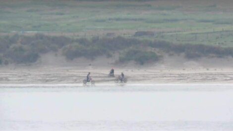 Three people ride bicycles along a sandy path by a body of water with grassy terrain in the background.