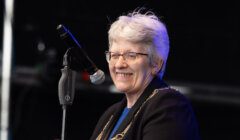 A smiling person with short white hair and glasses speaks into a microphone while wearing a black outfit with a ceremonial chain.