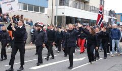 A group of people in navy uniforms march in a parade, some waving, with a Norwegian flag visible and a sign reading "Statsraad Lehmkuhl Norway." Spectators stand behind a low wall in the background.