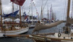 A row of boats with colorful flags docked at a marina during an event. Overcast sky in the background.