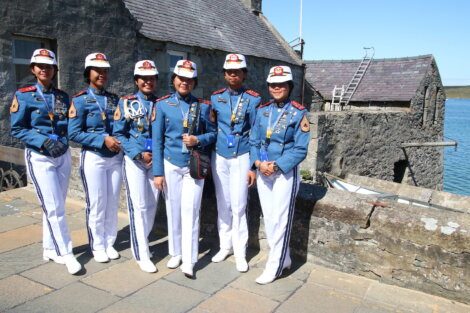A group of five people in blue military uniforms and white hats stand together outdoors near a stone building and water.