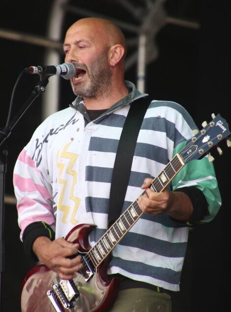 A person with a bald head and beard sings into a microphone while playing an electric guitar. They are wearing a colorful, striped long-sleeve shirt.