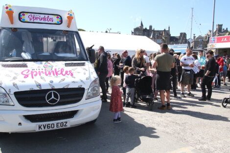 A crowd waits in line at an ice cream truck labeled "Miss Sprinkles" on a sunny day, with small children and adults gathered around. Tents and buildings are visible in the background.