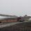 Heavy trucks transport large wind turbine blades on a gravel road in a foggy landscape. A service van is parked nearby.