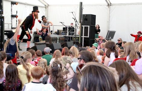 A performer dressed in a top hat and striped socks entertains a crowd of children and adults in a tented venue. Other performers and equipment are visible on stage.